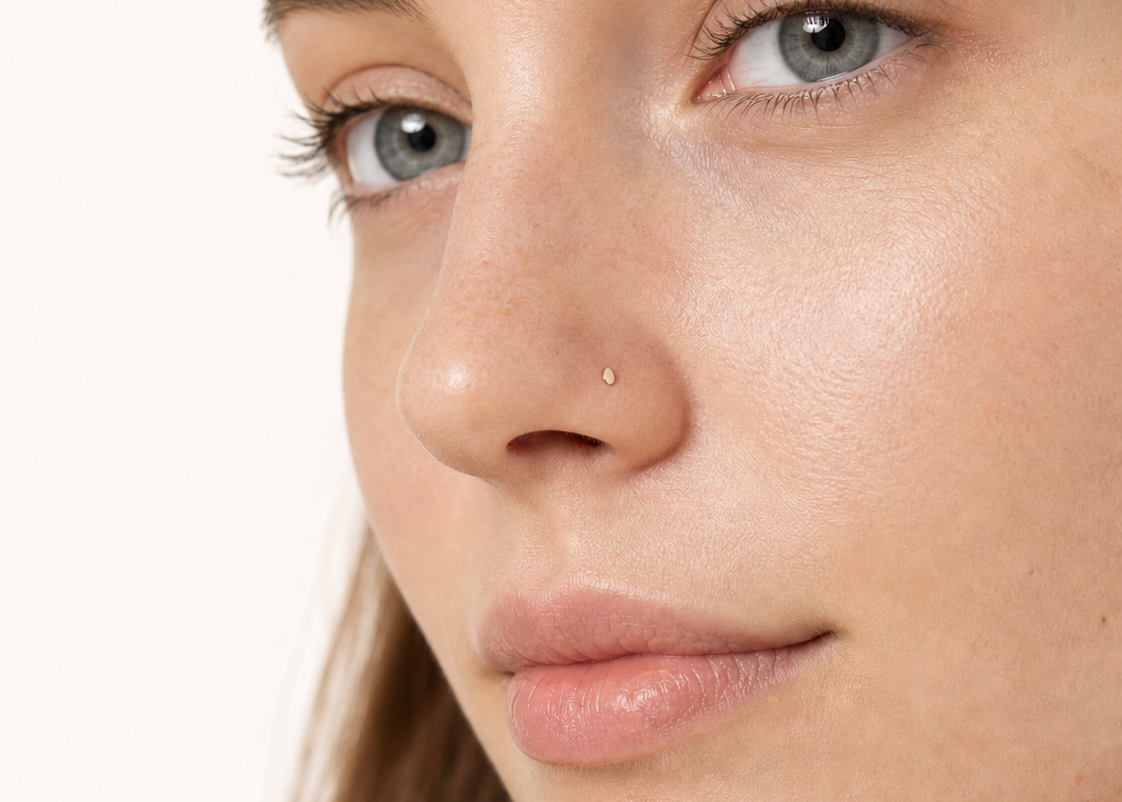 Close-up of a woman's face showing a tiny gold nose egg hoop piercing on her left side, against a clean off-white background.