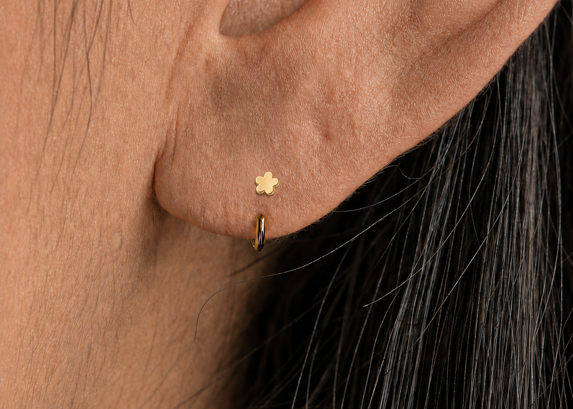 Close-up of a person's ear showing a small gold flower stud earring and a thin gold hoop earring.