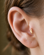 A small crescent moon gives the hoop a subtle celestial character with a polished solid-gold finish. Made for fully healed tragus piercings, it adds a compact decorative detail without turning the piece into a dangling design. Handmade to order in 14ct solid gold, it fits naturally with celestial, lunar and softly symbolic styling rather than reading as a generic plain hoop.