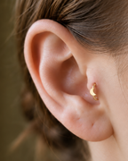 A small crescent moon gives the hoop a subtle celestial character with a polished solid-gold finish. Made for fully healed tragus piercings, it adds a compact decorative detail without turning the piece into a dangling design. Handmade to order in 14ct solid gold, it fits naturally with celestial, lunar and softly symbolic styling rather than reading as a generic plain hoop.