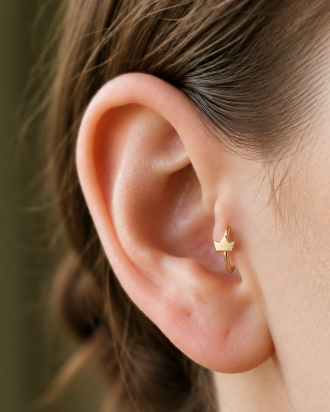 A tiny crown adds a playful statement detail while staying compact enough for everyday styling. Made for fully healed tragus piercings, it adds a compact decorative detail without turning the piece into a dangling design. Handmade to order in 14ct solid gold, it fits naturally with playful, symbolic and statement-led styling rather than reading as a generic plain hoop.