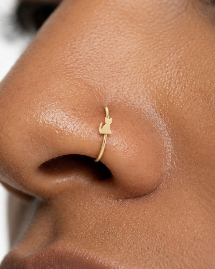 Keep a favourite animal close. This 14ct solid gold cat nostril ring pairs an approximately 3mm polished sitting-cat silhouette with a smooth seamless hoop. Pointed ears, a softly rounded body and an upright curved tail give the little cat its character.
Handmade to order in yellow, white or rose gold, with inner diameters from 6 to 10mm and a choice of 18G (1.0mm) or 20G (0.8mm). Both the cat and hoop are solid gold and nickel-free, designed for a fully healed nostril piercing.
Wear it alone for one playfu