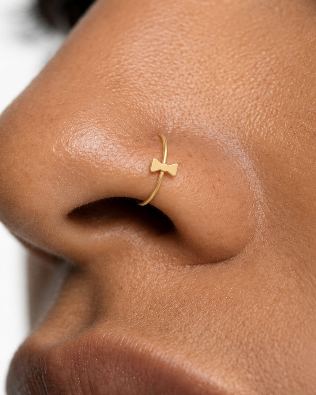 This miniature bow tie has two flared sides joined by a small central knot. Rounded outer corners soften its geometric outline, while the smooth gold surface keeps the design minimal. There are no trailing ribbon ends, engraved details or gemstones.

Handmade to order in nickel-free 14ct solid gold, this bow tie nostril hoop is available in yellow, white or rose gold. Choose a 6, 7, 8, 9 or 10mm internal diameter and 18G (1.0mm) or 20G (0.8mm). The motif measures approximately 3mm. Designed for fully healed
