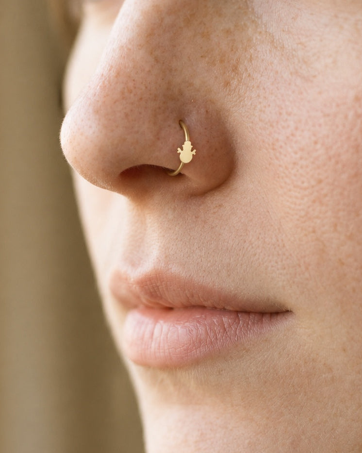 This miniature snowman wears a top hat and has a rounded body with two outstretched, twig-like arms. Its smooth gold surface has no engraved eyes, buttons or gemstones, keeping the winter silhouette simple and clear.

Handmade to order in nickel-free 14ct solid gold, this snowman nostril hoop is available in yellow, white or rose gold. Choose a 6, 7, 8, 9 or 10mm internal diameter and 18G (1.0mm) or 20G (0.8mm). The motif measures approximately 3mm. Designed for fully healed nostril piercings, with sizing c